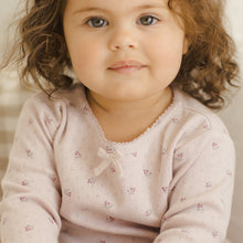 Load image into Gallery viewer, A young child with curly brown hair, dressed in the Pointelle Long Sleeve Tee + Legging featuring a lilac ditsy floral pattern and bow, sits indoors and looks calmly at the camera.