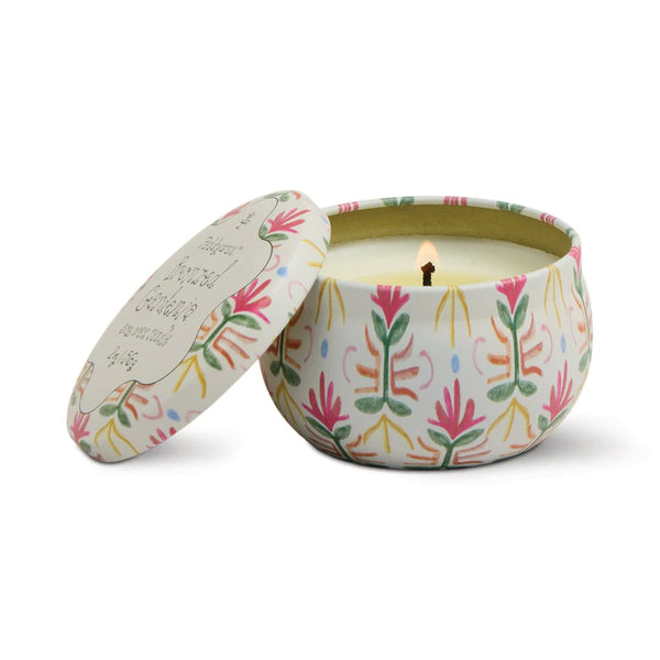 A lit Printed Tulip Candle Tin sits open, revealing its scented glow. The round tin features a decorative pink, green, and yellow floral pattern; the matching lid rests nearby.