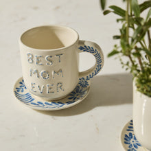 Load image into Gallery viewer, The Best Mom Mug/Saucer features a cream color with blue floral designs, making it an ideal Mother’s Day gift. A potted plant appears on the right side of a white countertop.