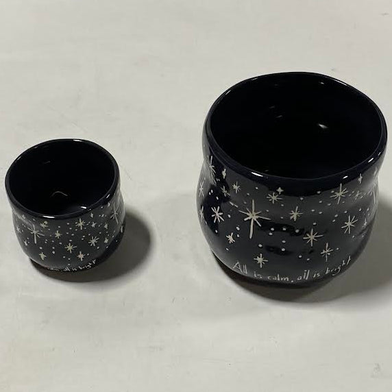 Two dark blue ceramic cups with white star patterns and “All is calm, all is bright” around the base, placed on a light surface. One cup is smaller than the other—perfect next to our Black Starry Planter or a holiday floral arrangement.