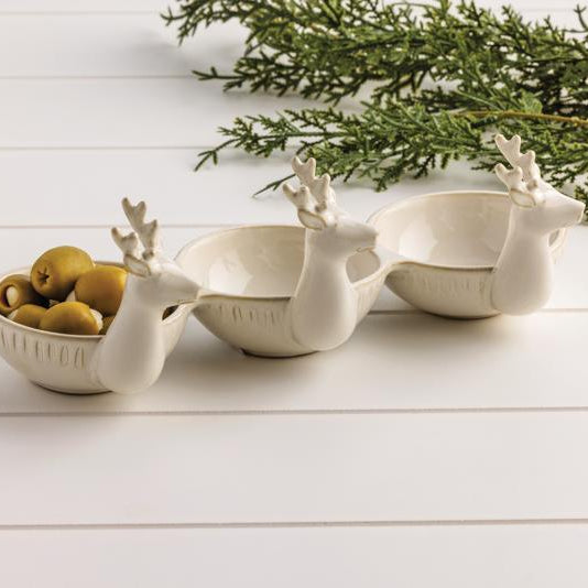 The Triple Deer Tidbit features three white ceramic dishes shaped like reindeer heads, arranged in a row. One holds green olives, while greenery and a white wood surface create a festive holiday setting.