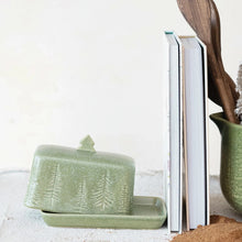 Load image into Gallery viewer, The Green Debossed Stoneware butter dish, featuring a tree design and a tree-shaped handle, sits next to upright books and a green mug with wooden utensils against a neutral background.