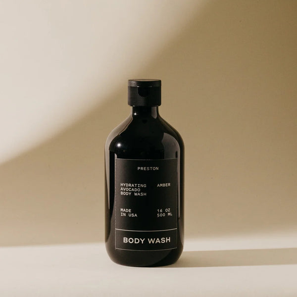 A dark bottle of Preston Body Wash stands on a light surface with a beige background. The label describes it as a moisturizing wash with hydrating avocado oil, made in the USA, and containing 16 oz (500 ml).