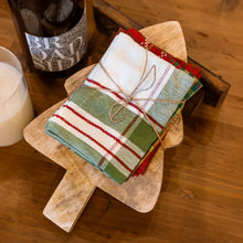 Load image into Gallery viewer, A stack of colorful plaid napkins tied with twine sits on the Tree Board Gift Set—a Mango Wood Christmas Tree Cutting Board. A glass of milk and a bottle nearby add festive charm to your kitchen decor.