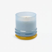 Load image into Gallery viewer, The 7oz Stacked Glass Candle, made with white soy wax, glows in a translucent, multi-colored glass holder with a rounded base featuring yellow and blue accents, set against a plain white background.