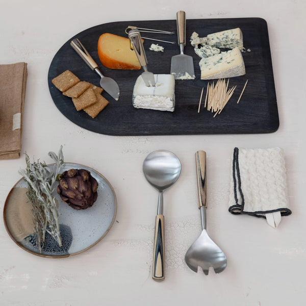 A cheese board with assorted cheeses, crackers, and toothpicks sits beside a plate with a purple artichoke and herbs. Three metal serving utensils and a folded S/3 Dish Cloths w/ Colored Edge are also arranged on the table.