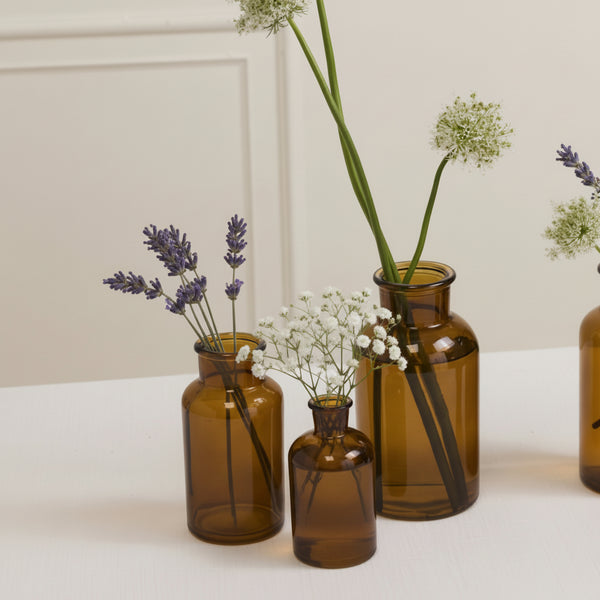 Three Transluscent Amber Bud Vases, each filled with lavender and white baby's breath, are arranged elegantly as budvases on a white surface against a cream-colored wall.