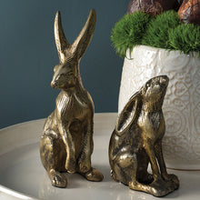Load image into Gallery viewer, Two gold Gold Rabbit figurines with elongated ears, one upright and one looking up, sit on a white tray. Behind them, Easter decor is enhanced by a decorative white pot filled with green moss and brown accents.