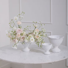 Load image into Gallery viewer, A modern white marble table features a floral vase with pale pink and white flowers, a Rustic White Compote, and two empty ceramic bowls of varying sizes, all set before a light paneled wall.