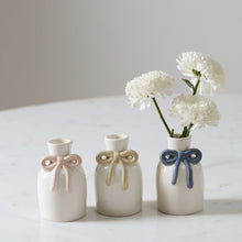 Load image into Gallery viewer, The Budvase w/ Bow set features three small white ceramic vases with pastel pink, yellow, and blue bows—perfect for spring event decor. The vase with the blue bow displays white flowers while the others sit empty on a marble surface.