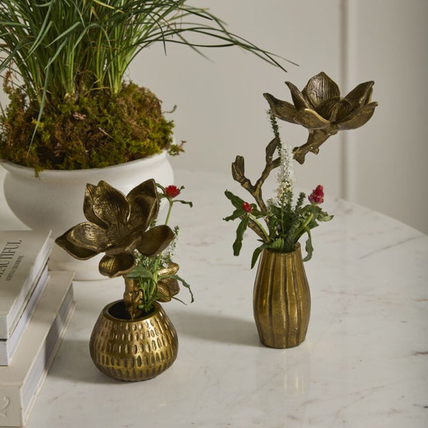 Two Golden Cast Aluminum Flower & Pot pieces with metal floral designs and small flowers sit on a white marble table, creating a charming spring display beside a stack of books and a white planter with grass and moss.