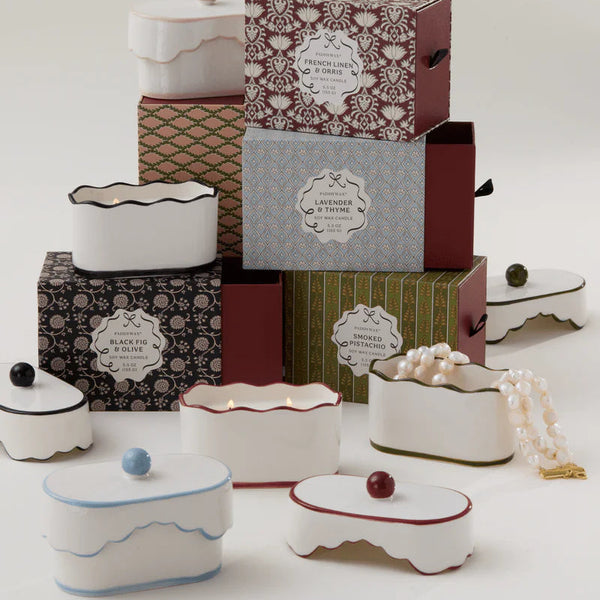 The 5.5oz Ceramic Dish Candle by Paddywax in white lidded containers is displayed with boxes labeled lavender, thyme, and black fig; a pearl necklace rests nearby.