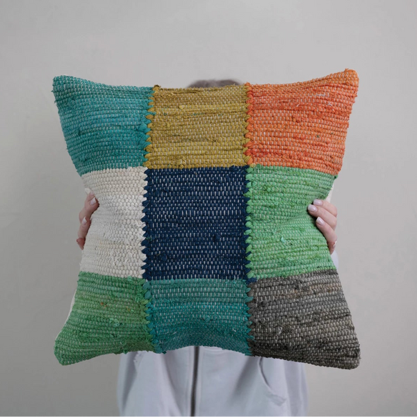 A person in a white top holds up the 20" Multi Color Checked Pillow, which features a vibrant patchwork of green, blue, yellow, orange, gray, and cream squares against a plain light background.