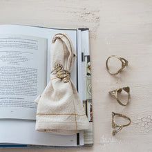 Load image into Gallery viewer, A set of four Brass Leaf Napkin Rings is displayed, with one on a beige napkin atop an open cookbook and the other three—each featuring distinct leaf designs—arranged vertically on a textured white surface beside the book.