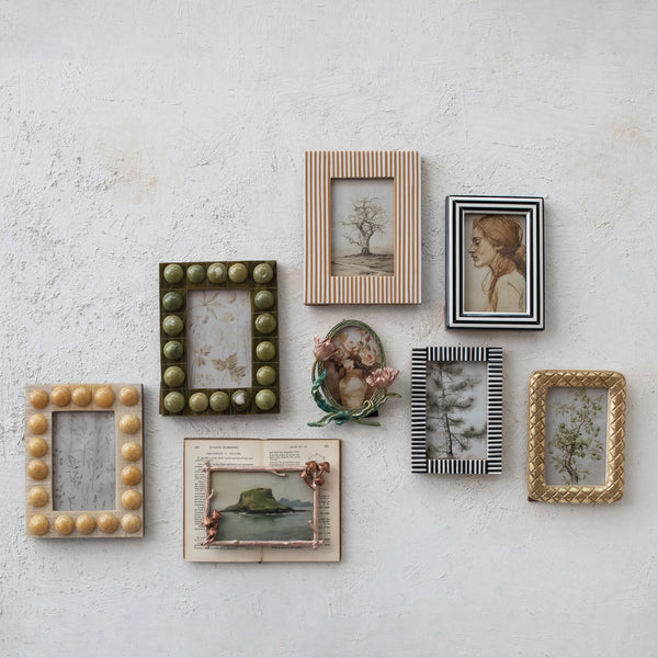 Seven framed artworks are arranged on a textured white wall, featuring paintings, botanical prints, and an illustrated open book in decorative frames such as an elegant frame and a 4x6 Resin Striped Frame.