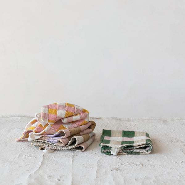 A stack of folded pastel checked and plaid cotton Dish Cloths w/ 4 Checked & Plaid Styles sits on the left, next to two green and white checkered cloths, all placed on a light textured surface with a plain white background.