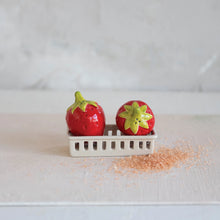 Load image into Gallery viewer, The Strawberry S+P Shakers, shaped like ceramic strawberries, rest in a small berry basket holder on a light surface with pink granules scattered nearby and a plain, light background.