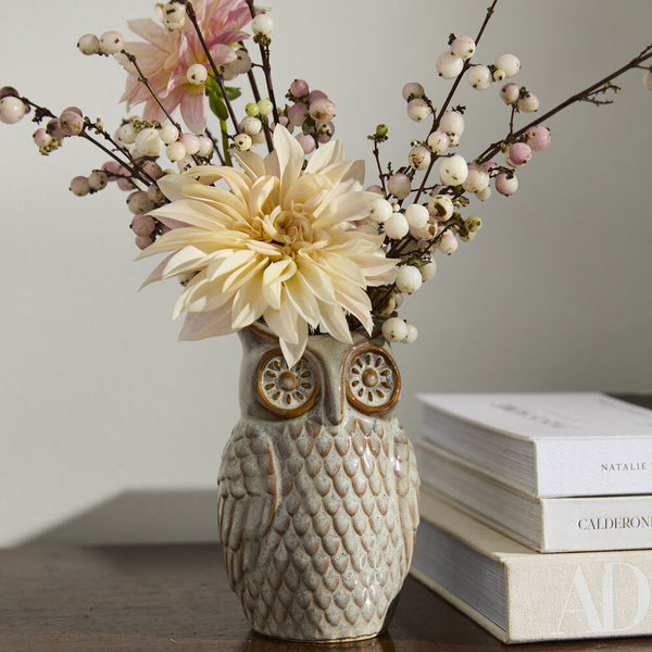 The Great Horned Owl Vase, featuring a grey reactive glaze, displays cream dahlias with white and pink berry sprigs on a wood surface next to stacked books.
