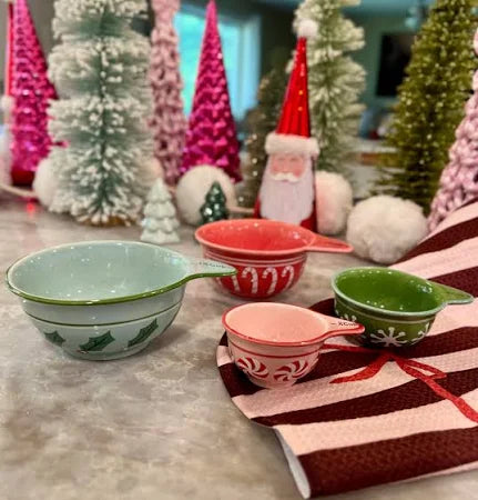 Christmas Stoneware Measuring Cups feature festive holiday designs and are arranged on a counter next to a red and white towel, with colorful bottle brush trees and a Santa figurine in the background—perfect for holiday baking.
