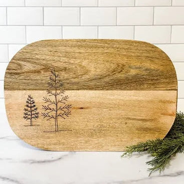 The Mango Wood Tree Cutting Board features two engraved pine trees on the left side, rounded edges, and is shown resting on marble with green foliage and a white subway tile background.
