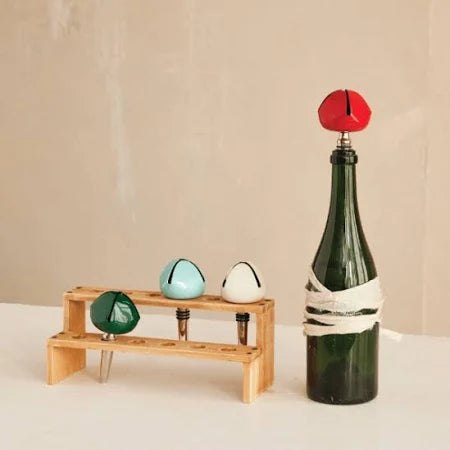 The Bell Bottle Stopper, in green glass with a metal top, sits beside a wooden rack displaying three stoppers—green, light blue, and white—offering a stylish wine gift. Set against a plain beige background.