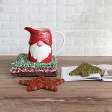 Load image into Gallery viewer, A ceramic gnome-shaped pitcher rests on a Holiday 8&quot; Crocheted Pot Holder beside an open book, with green tree and red star coasters on a wooden table. A white brick wall forms the background.