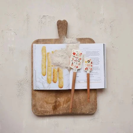 A cookbook open to a recipe page lies on a wooden cutting board. The Set/2 Christmas Spatula, both flour-dusted with festive handles, rest atop the book—perfect for your holiday baking—with flour scattered on pages and board.