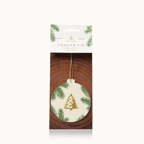 The Frasier Fir Heritage Collection sachet features festive ornament-shaped packaging with green pine branches and a gold tree design, delivering a refreshing winter fragrance against a wood-patterned background.