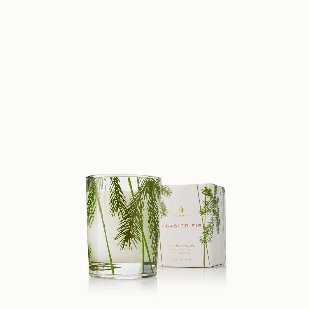 A Frasier Fir Heritage Collection glass candle, adorned with green pine needle designs, sits beside its matching box against a light background, capturing the essence of an evergreen forest.