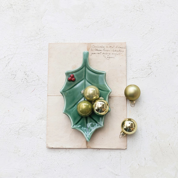 A Holly Leaf Dish with a reactive glaze displays gold Christmas ornaments atop a vintage letter on a light textured background, with three additional gold ornaments placed beside the dish.