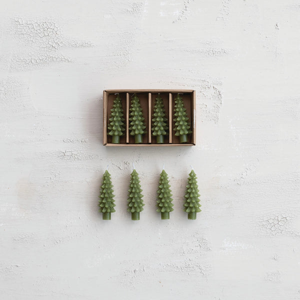 Four green, unscented pine tree-shaped candles from the Box/4 Shaped Candles set sit in a brown box, with one displayed outside—all shown on a textured white surface, ideal for holiday décor.