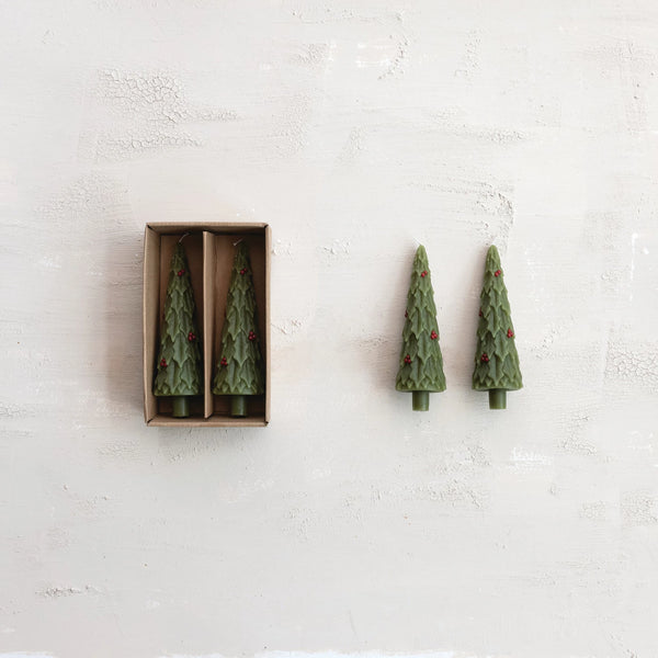 The S/2 Taper Tree Candles w/Holly—two green, unscented tree-shaped tapers with red holly decor—come in an open box and rest on a textured surface, adding festive charm to your holiday décor.